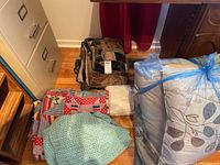 Lot overview showing vintage quilt, pillows, packaged blanket, and other bedding items on wooden floor near filing cabinet and desk