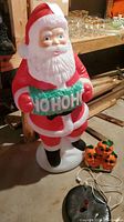 Photo showing a blow mold Santa figurine holding a green sign with 'HO HO HO' text and a small blow mold pumpkin decoration beside it on a black circular base.