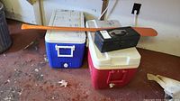 Two large coolers, one blue with white lid and one red with white lid, on the floor with wooden canoe paddle laid across and black tackle box on top of red cooler.