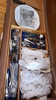 Drawer open displaying silver plated cutlery including spoons, tongs, and other serving utensils wrapped partially in tissue.