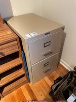 Overall view of the beige Commodore two drawer filing cabinet placed beside a wooden shelving unit on a hardwood floor.