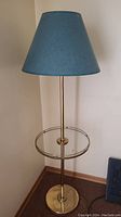 Full view of the tri-light floor lamp showing the brass finish, circular glass table, and teal fabric lampshade.