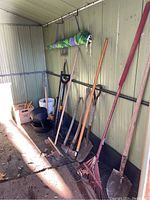 Photo of garden tools including shovels, a rake, axes, and other tools leaning against a green painted wooden wall in a shed.