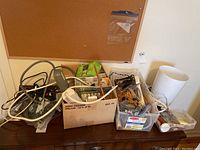Wide view showing two boxes and one transparent storage bin filled with power cords, adapters, electrical tape, a lamp shade, and other household items.