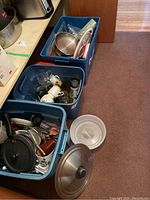 Three blue plastic bins on floor containing assorted kitchen items including metal pots and pans, glassware, and utensils.