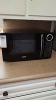 Black RCA microwave oven on a kitchen countertop, showing front and digital control panel.