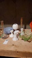 Full view of lot items showing the white milk glass candle holders, green glass bowl, blue glass bowl, a white hobnail vase, and two tall glass vases with gold designs on wooden surface.