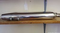 Single chrome motorcycle muffler lying horizontally on wooden shelf with two mounting clamps attached.