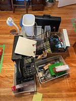 Full view of office supplies lot including briefcase, phone, file folders, plastic bins with office tools, desk lamp parts, and trash bin on wood floor.