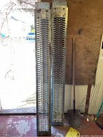 Two metal truck ramps standing vertically against a wall near a shovel, showing their grated tread surface and bolt holes on top plate.