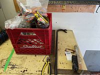 View of crate filled with hardware items and tools, yellow-handled pliers, crowbar, wire brush, and flat metal bar on workbench surface.