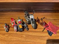 Five toy motorcycles lined up on a wooden floor along with two small miniature motorcycle bags, one red and one black with red accents.