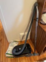 Black Kenmore canister vacuum with grey hose and metal wand standing on wood floor next to unopened vacuum bags.