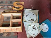 View of three floral china wall plates placed next to a wooden wall rack and a decorative wooden letter 'S'. The plates are white with floral designs and holes for hanging.