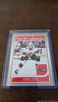 Photo of the Shane Pinto O-Pee-Chee rookie hockey card in a clear protective sleeve on a wooden surface.