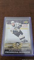 Photo of Sidney Crosby rookie hockey card in a protective sleeve featuring Crosby skating in a white Pittsburgh Penguins uniform with his name and card number visible.