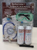 Front view of Duracool 22a refrigerant recharge kit sealed in plastic packaging showing two refrigerant cans, pressure gauge, hose, valve tool, and goggles.