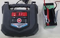 Front view of Schumacher SC1323 battery charger with digital display showing 3.3 amp output, main control panel with Start/Stop button and attached charging clamps on a holder.