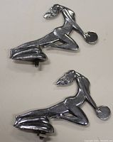 Top view of two silver goddess hood ornaments showing front profile, figure kneeling with long flowing hair and circular disks in their hands.