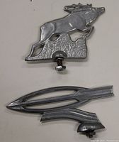Pair of two metal hood ornaments placed side by side on a flat surface showing front and side views.