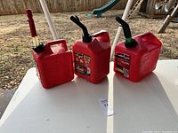 Photo of three red plastic gas cans on a white table outdoors, showing the red pump handle and flexible black spouts with green rings.