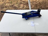 Photo of the full blue hydraulic trolley car jack with long lifting handle displayed on a white fold-out table outdoors.