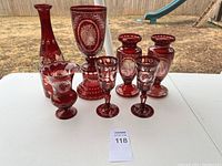 Overview of eight ruby red glass items with white etched floral motifs and scrollwork on white table outdoors.
