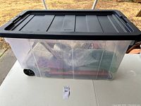 Clear large plastic storage tote with black lid and wheels containing assorted tools and materials such as foam pieces and fabric