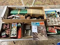 Overview of all vintage model train buildings, accessories, train cars, and magazine packed inside cardboard boxes on a table.