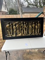 Front view showing ten elongated abstract human figures in gold relief on a black backing within a black wooden frame.