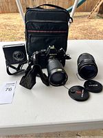 Nikon N2000 camera body with attached lens, external flash, extra zoom lens with lens caps, and black carrying bag on white table outdoors.