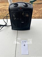 Front view of Pelonis small black electric heater on white table outdoors with control knobs and power cord visible.