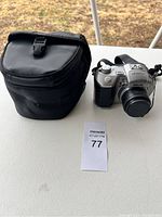 Front view of the Sony camera with lens cap next to the black padded carrying bag on a white surface outdoors.