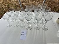 Wide view of all 15 vintage etched glasses on white table outdoors showing two sizes of glassware with floral etching.