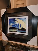 Framed fine art print showing stylized mountains, lake, and sky, protected by glass, with black wooden frame and white-lined black mats.