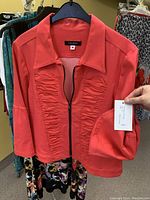 Coral red women's jacket from VALERIE with ruched detailing and zipper, held by hand showing tag