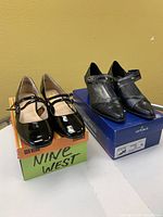Two pairs of ladies shoes on their original boxes, one pair black patent leather Mary Jane style, one pair black leather Mary Jane style with pointed toe and ankle strap.