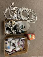 Top-down view of two cardboard bins showing white electrical cords, a white lamp shade, several lightbulbs both packaged and loose, and various small electrical components.