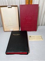 Photo showing the black Holy Bible with gold lettering, the protective box, red outer box with embossed tree logo and guarantee card.