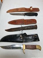 Three vintage cutting knives with their leather sheaths laid out on a white surface, showing wear and usage.