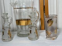 Overview of the full set including the pitcher, two oil and vinegar containers, and tall drinking glass. Shows gold leaf decoration and gold banding on the pitcher and glass.