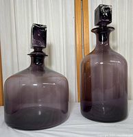 Two large purple glass containers with wide bases and square glass stoppers, placed side by side on white cloth against wood and ribbed white background.