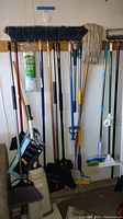 Photo showing a variety of cleaning tools including brooms with plastic bristles, mop, and vacuum cleaner in a garage setting.