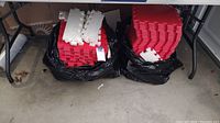 Two large black bags filled with red and white interlocking foam floor mats stacked under a table in a garage.