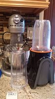 KitchenAid Professional HD stand mixer shown on counter with stainless steel bowl and wire whisk inside, alongside Omega smoothie maker with blending containers attached.