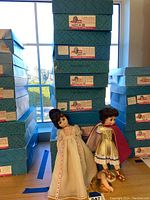 Two standing dolls in costume showing one with detached arm and shield, surrounded by stacks of blue boxes.