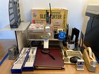 Wide view of desk containing assorted office supplies including stapler, tape dispenser, desk paper sorter, pens, magnifying glass, and Villeroy & Boch letter openers in boxes.