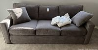 Full view of the gray velour fabric three-seater sleeper sofa bed with two throw pillows and linens on the seat.
