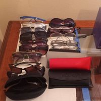 Top-down view of ten pairs of glasses arranged in two rows on a wooden table, including sunglasses and clear lens reading glasses, along with three glasses cases at the bottom.