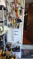 Wide view of pegboard with assorted hand tools including pliers, hammer, broom and other hardware items hanging; also reveals part of shelving unit and entry door.